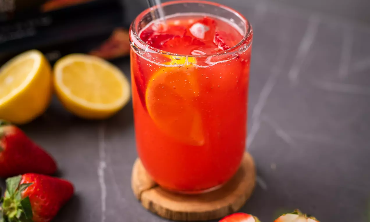 Dzidrs stikla glāze ar sarkanu dzērienu, peldējošu citrona šķēli un ledus gabaliņiem, kas novietota uz koka paliktņa, blakus svaigām zemenēm