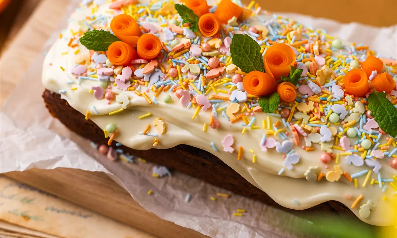 Garena, siltā oranžā tonī cepta mīksta morku keksas virsū bagātīgs baltā glazūras klājums, dekorēts ar konfektēm, burkānu rullīšiem un svaigām piparmētru lapiņām