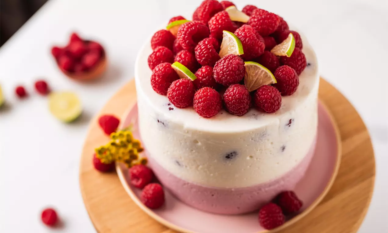 Divu slāņu torte ar viegli rozā un baltu krēmu, bagātīgi dekorēta svaigām aveņu kaudzēm un laima šķēlītēm uz koka paplātes.