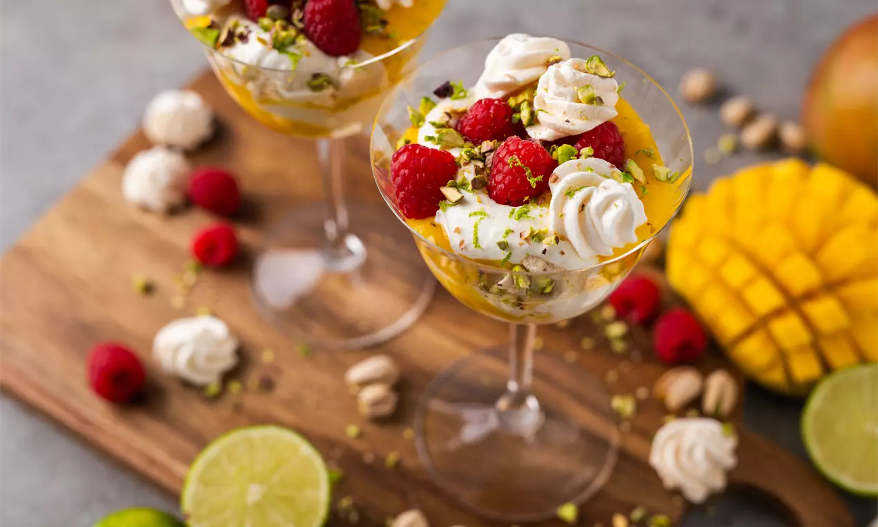 Stikla desertu glāzēs gaisīgs krēms ar dzeltējošu mango mērci, rotāts ar avenēm, pistāciju gabaliņiem un mazām morengu rozītēm